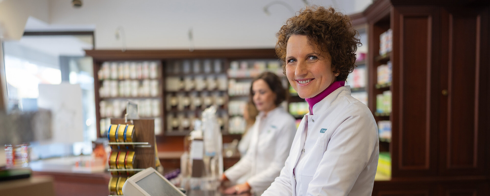 Apothekerin blickt lächelnd zur Kamera, im Hintergrund arbeitet eine Kollegin im Verkaufsraum einer gut sortierten Apotheke.