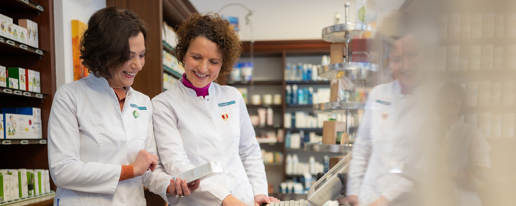 Zwei Apothekerinnen im Gespräch, betrachten gemeinsam ein Tablet in einem hellen Apothekenraum mit Medikamentenregalen.