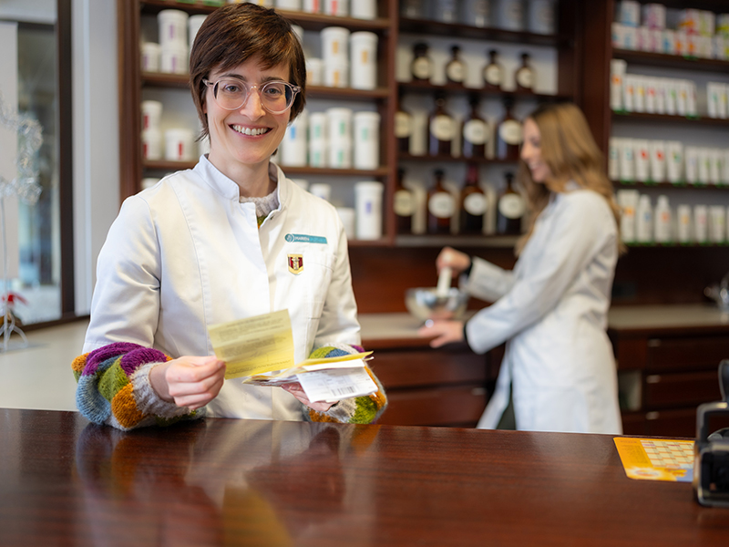 Apothekerin steht mit Rezepten an der Offizintheke, im Hintergrund wird mit Mörser gearbeitet – klassische Apothekenszene.
