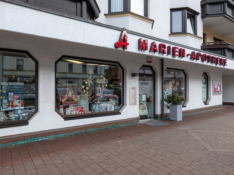 Frontansicht der Marien-Apotheke mit großen Schaufenstern, rotem Schriftzug und dekoriertem Eingangsbereich bei bewölktem Wetter.
